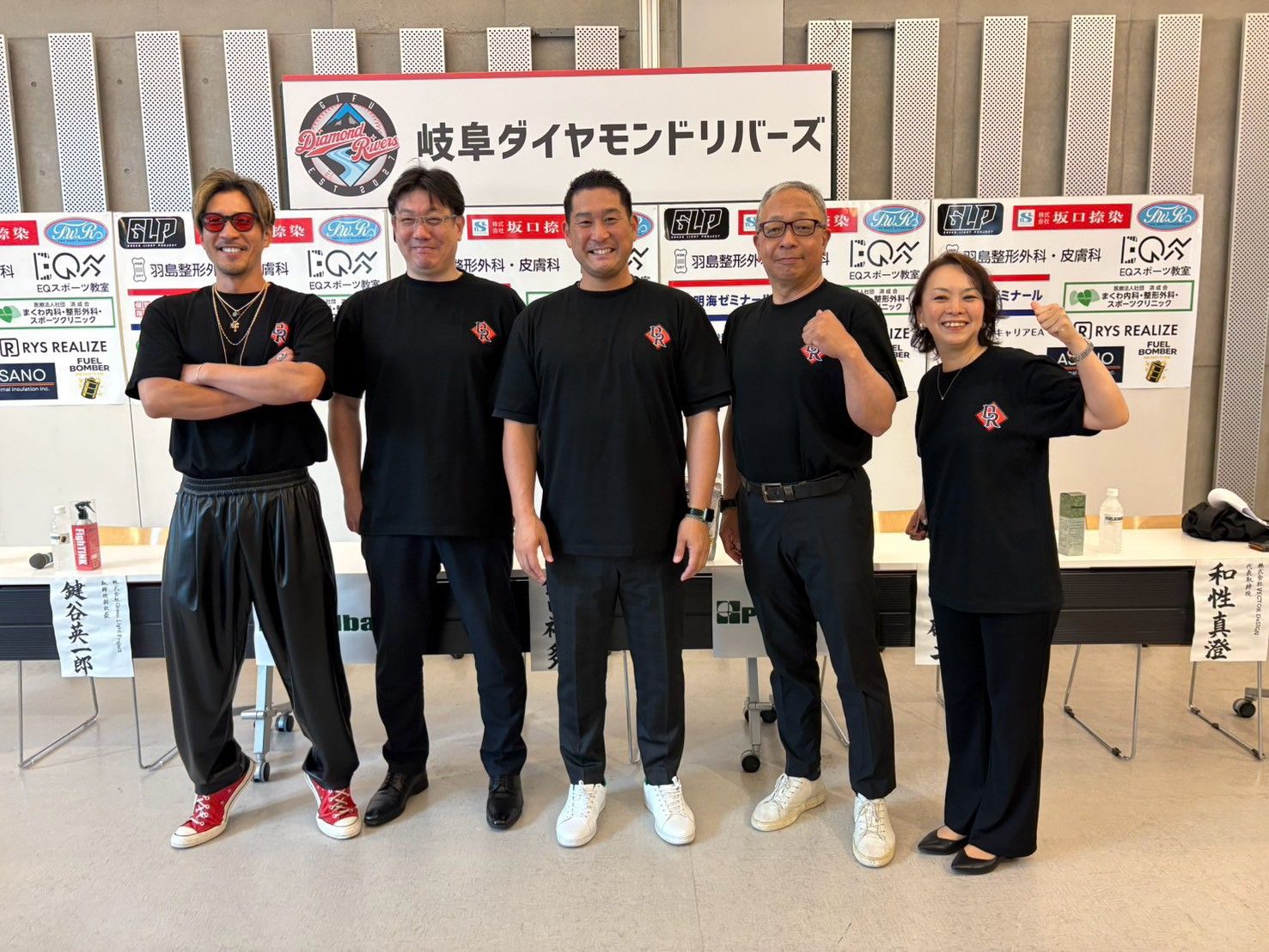 球団名入りの看板とスポンサーボードの前で、黒い球団Tシャツ姿の球団関係者5人が拳を握って並ぶ記者会見の様子
