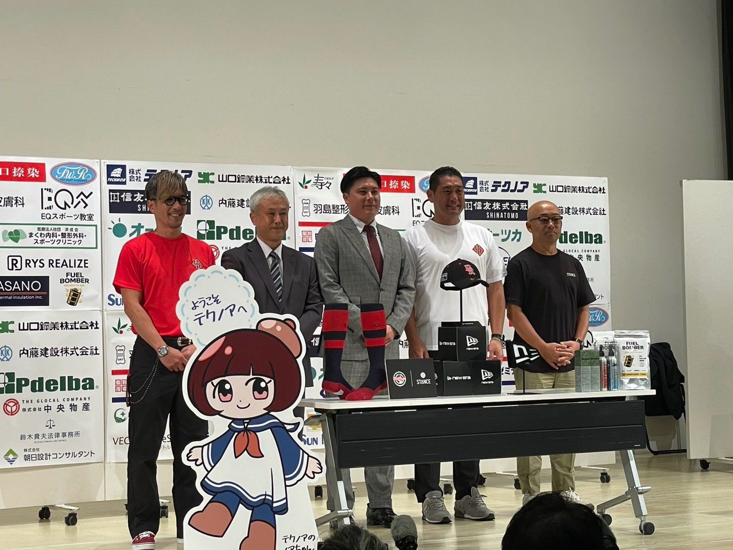 就任会見のステージに並ぶ登壇者5人。中央にグレーのスーツの若松監督、机にはキャップやソックスが並ぶ