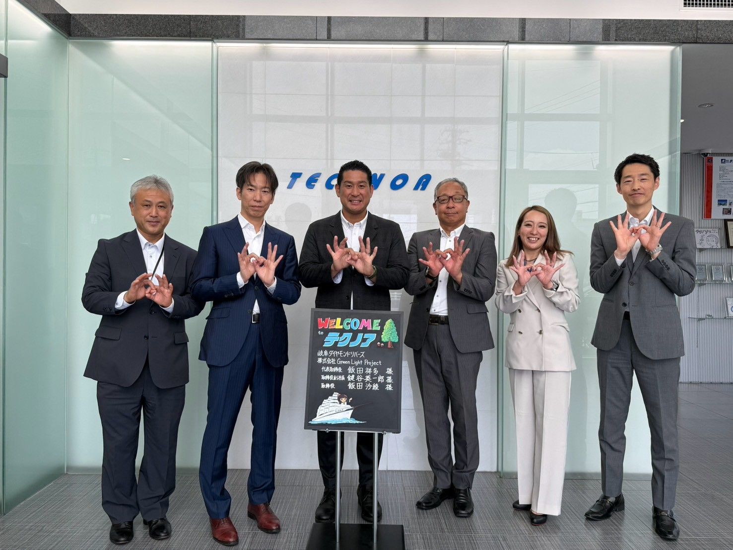 TECHNOAのロゴがあるエントランスで、歓迎ボードを囲んで並ぶ6人